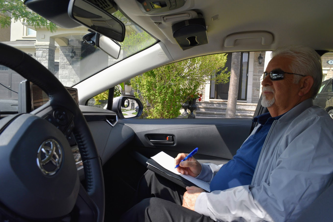 Nadi providing professional driving instruction, taking notes on clipboard while supervising a student driver from the passenger seat