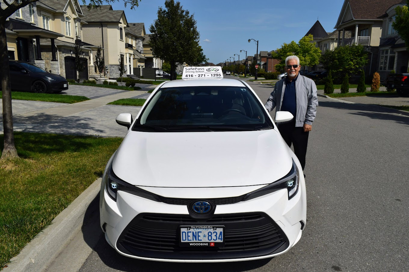 Nadi, SafePass Driving School instructor, standing next to the white Toyota Corolla used for driving lessons with SafePass sign on roof